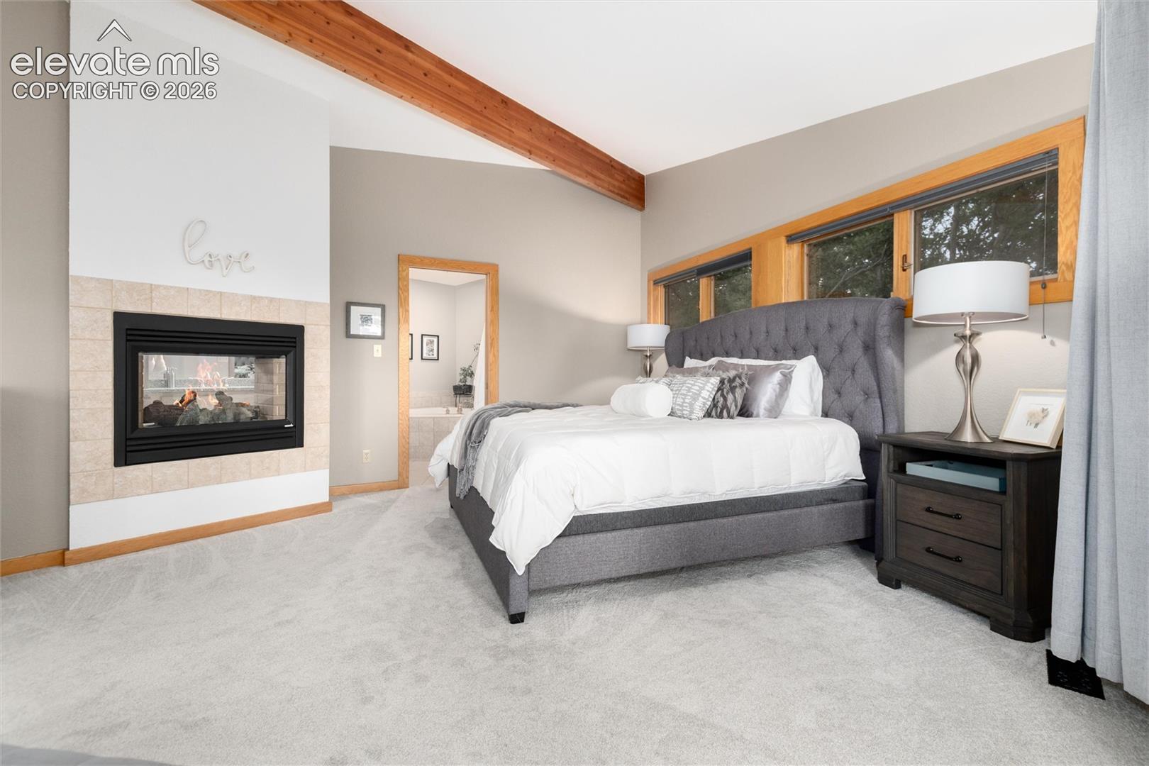 Room featuring a gas fireplace with a tiled surround, exposed wood beams, and warm wood-framed windows