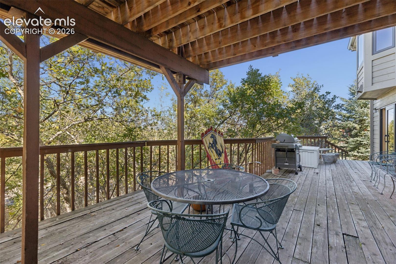 Covered wooden deck featuring an exposed beam ceiling, wooden railings, and mature tree surroundings