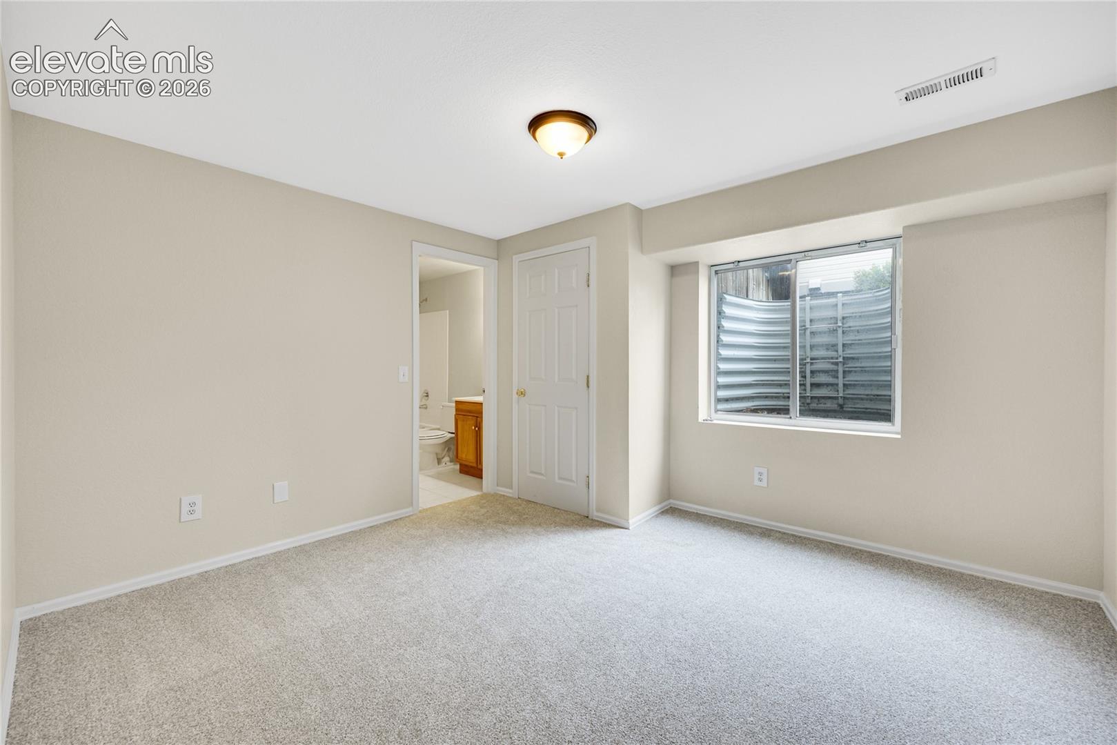 Neutral-toned room featuring neutral carpeting, a semi-flush mount ceiling fixture, and a window with a corrugated metal egress well