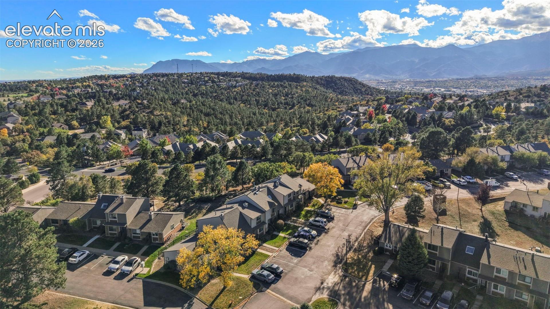 Aerial perspective featuring multi-level residential structures, paved parking areas, mature deciduous and coniferous trees, and a mountain range backdrop