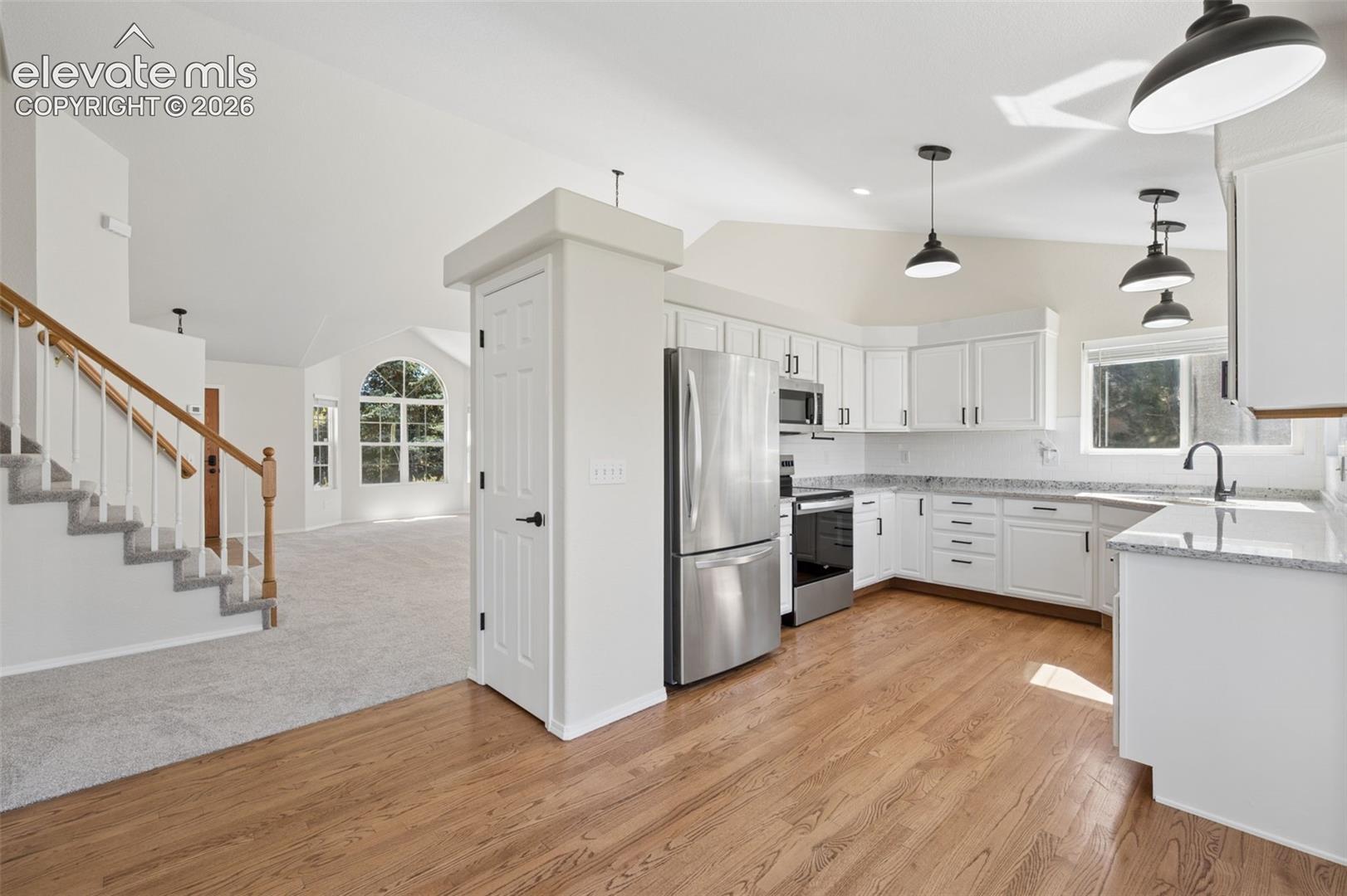 Kitchen featuring white cabinetry, stone-finish countertops, stainless steel appliances, and pendant lighting, set against wood-finish flooring and a staircase with a wooden handrail