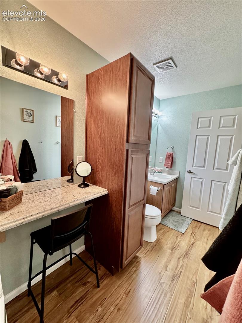 Bathroom featuring wood-finish flooring, a vanity with a light-colored countertop and integrated sink, a separate makeup vanity with a similar countertop, and a tall wood-finish storage cabinet