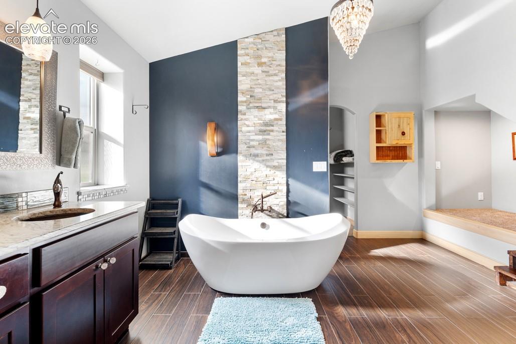 Freestanding soaking tub with dark wood-finish flooring, a stacked stone accent wall, and a crystal chandelier