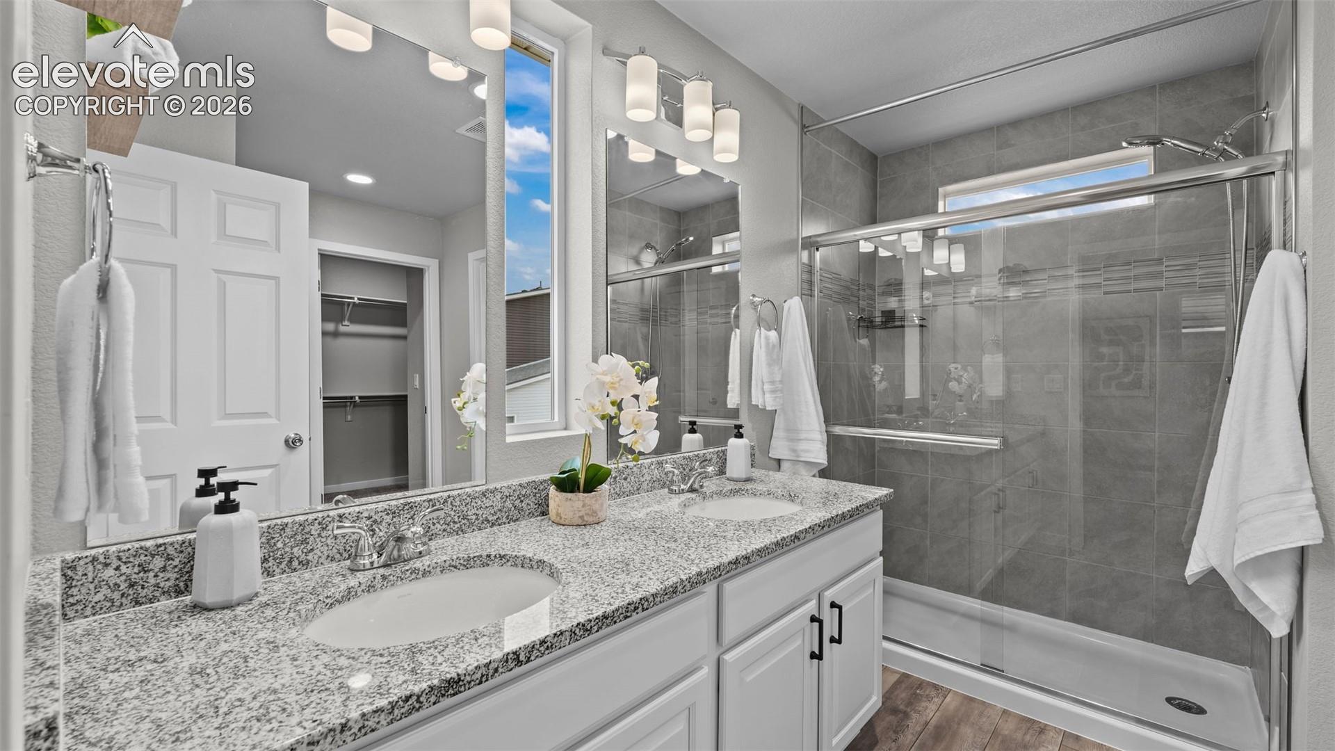 Dual-sink vanity with stone-style countertops, shaker-style cabinetry, glass-enclosed shower featuring tiled surround, and wood-finish flooring