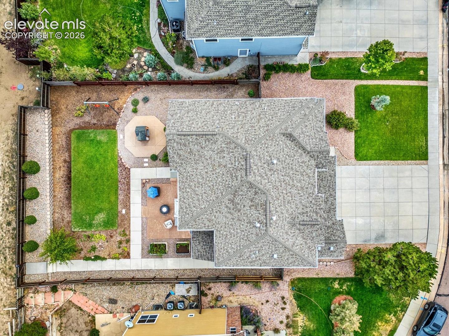 Aerial perspective of a property featuring a concrete driveway, multi-section asphalt shingle roof, paved patio area, maintained lawn, and fenced perimeter