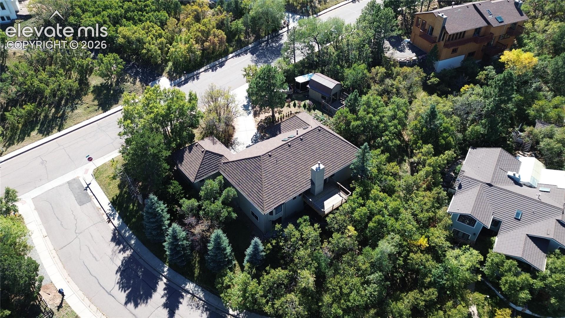 Aerial perspective of a multi-level residence featuring a concrete tile roof, elevated rear deck, exterior masonry chimney, and a concrete driveway situated within a heavily wooded lot