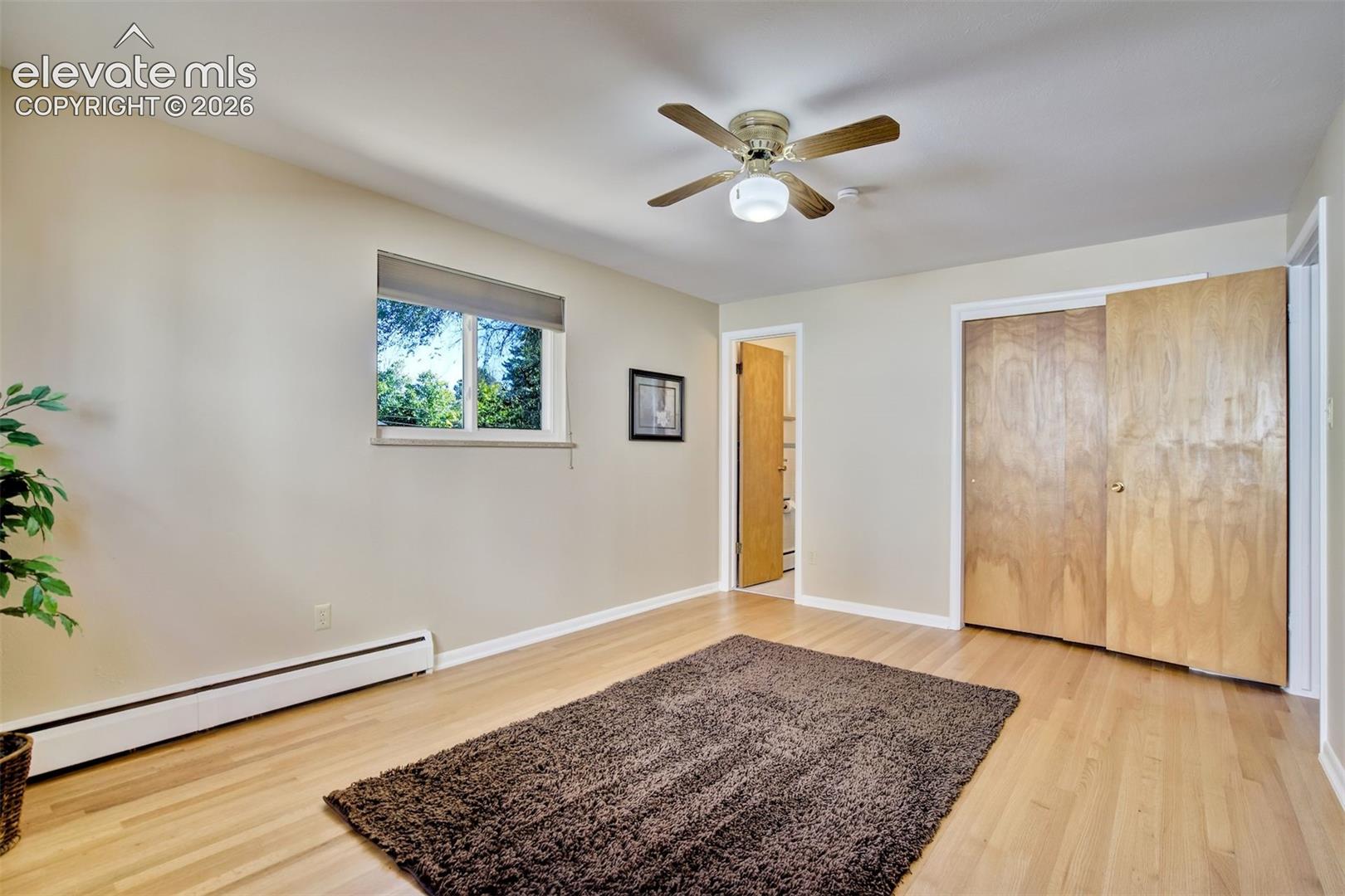 Room with wood-finish flooring, ceiling fan with integrated light fixture, natural wood-finish closet doors, and single-pane window with roller shade