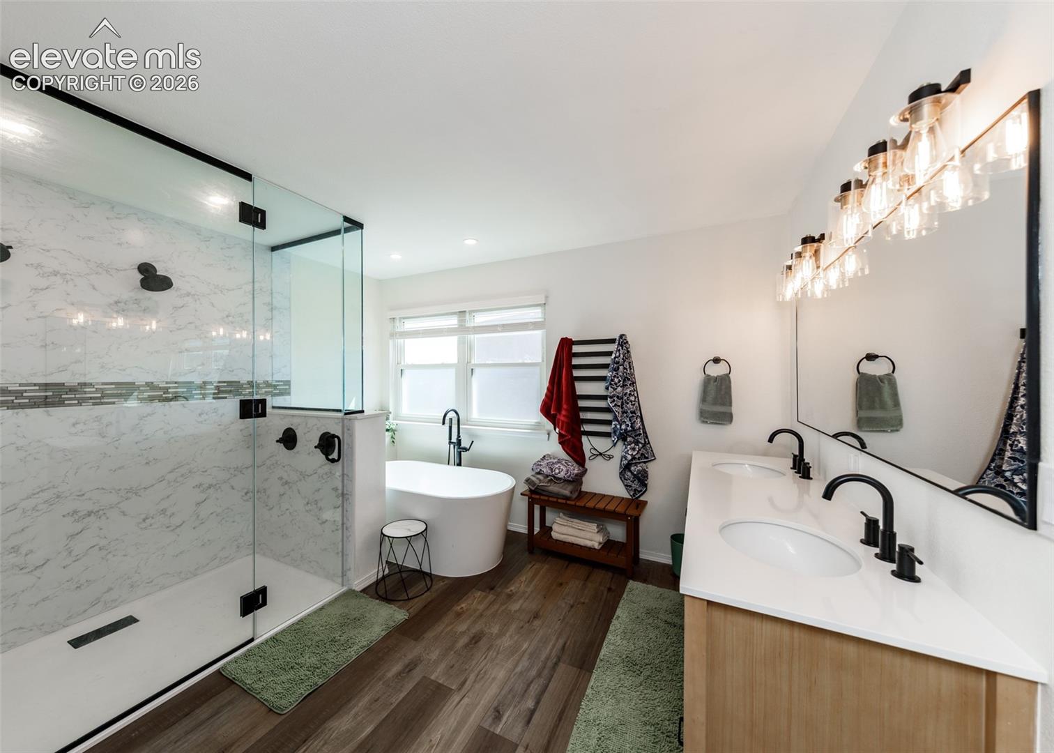 Spacious bathroom featuring a glass-enclosed shower with tile surround, freestanding soaking tub, dual vanity with undermount sinks, and wood-finish flooring
