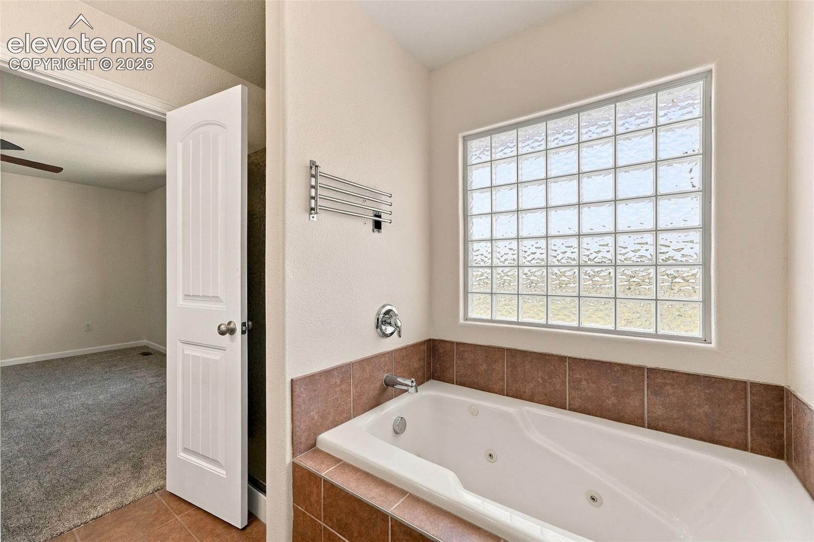 Jetted soaking tub with tile surround, glass block privacy window, chrome plumbing fixtures, and metal wall-mounted towel rack