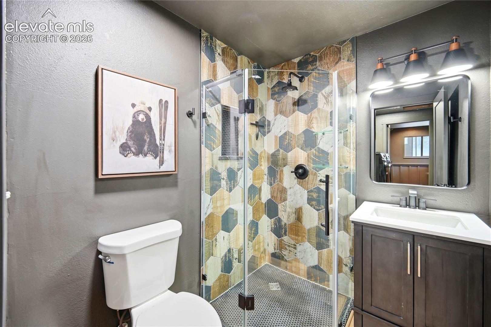 Corner shower enclosure with multi-toned hexagonal tile surround, dark wood-finish vanity with white countertop, and three-light wall-mounted fixture