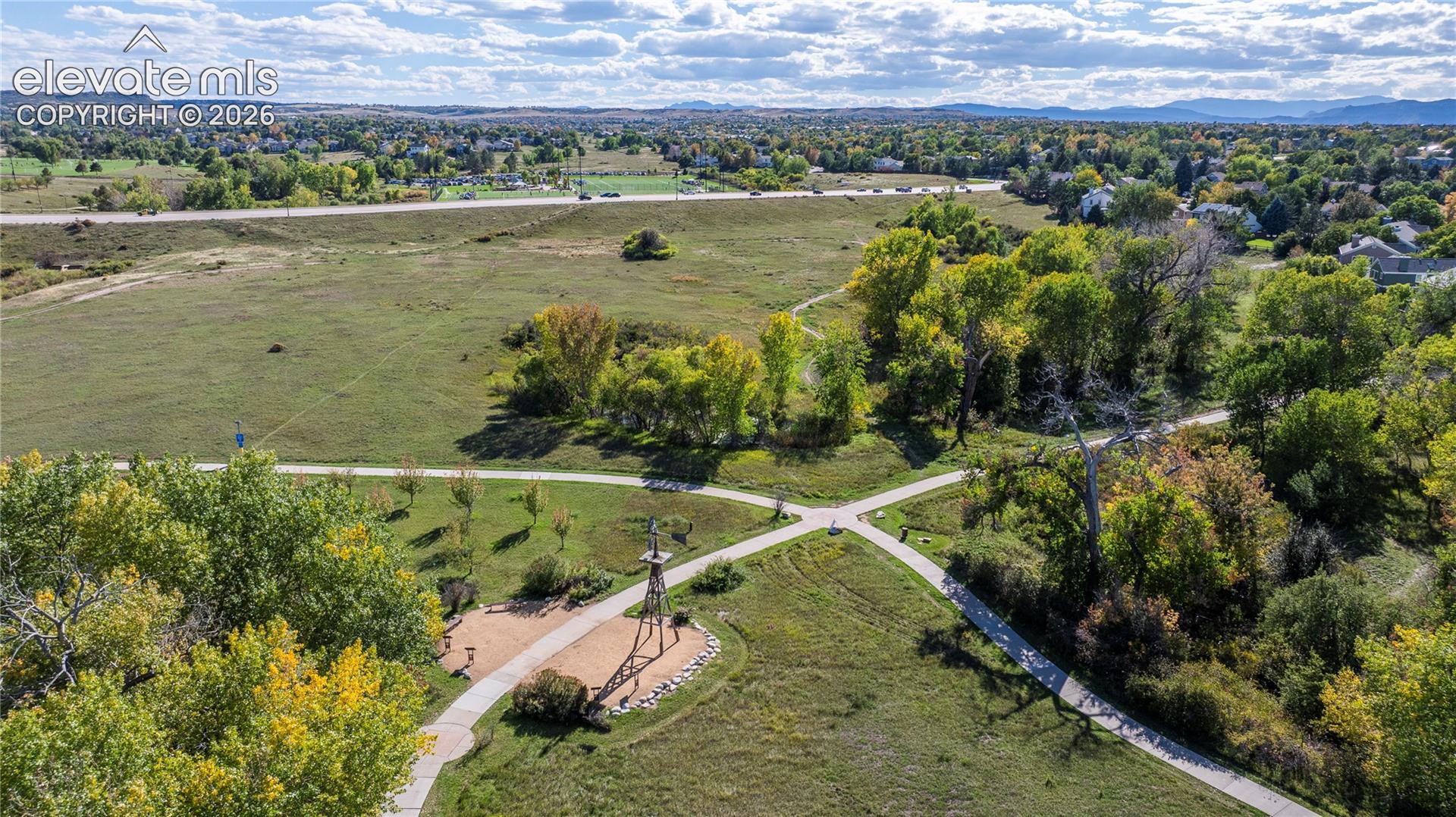 Aerial perspective of expansive open space featuring paved pedestrian pathways, clusters of mature deciduous trees, a central metal structure, and distant mountain topography