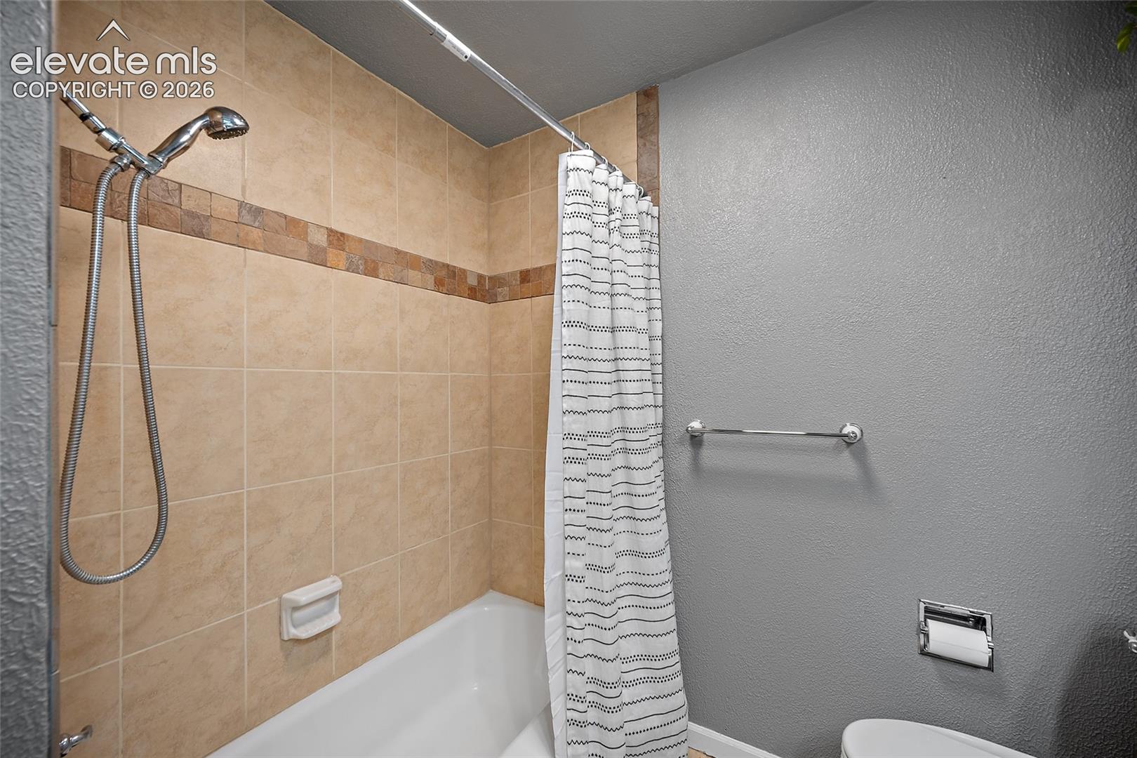 Bathtub enclosure featuring beige square wall tiles with a decorative mosaic accent border, handheld shower fixture, built-in soap dish, and wall-mounted metal towel rack