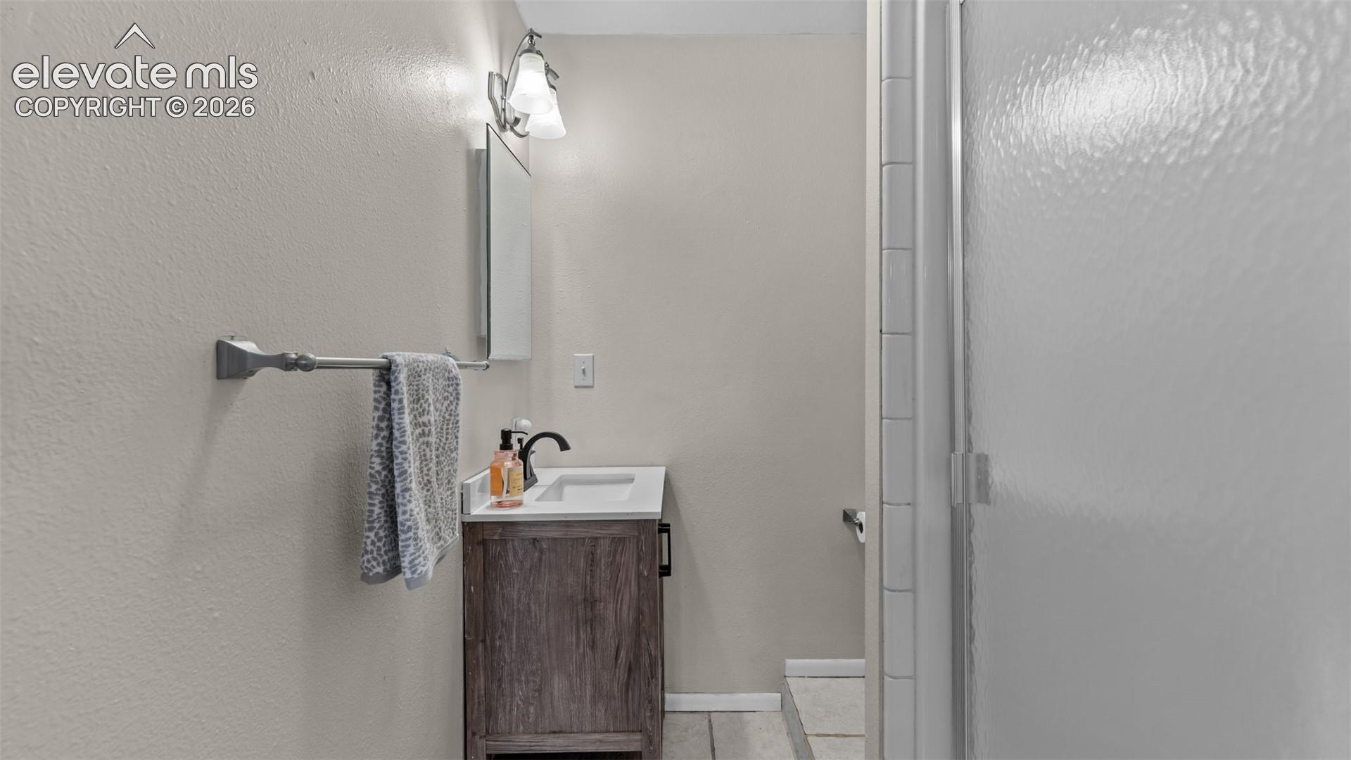 Single-sink vanity with a stone-top basin, wood-finish cabinetry, dark metal hardware, wall-mounted vanity light, and a shower with glass enclosure