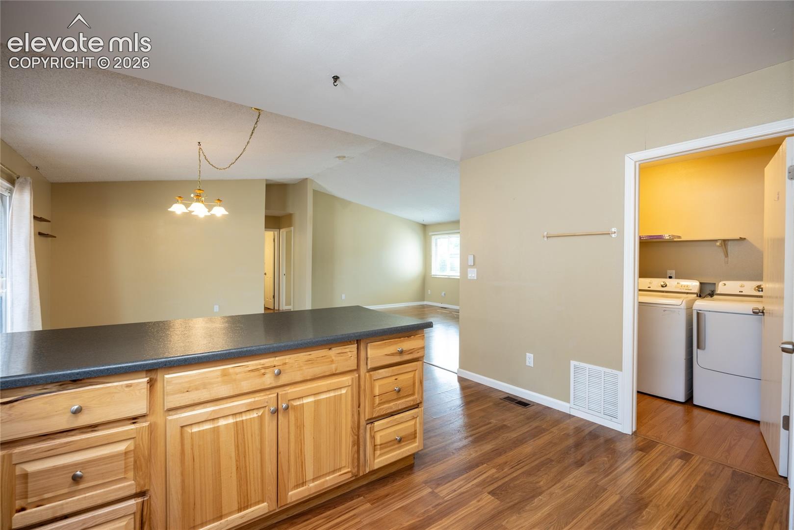 Kitchen island with wood cabinetry and dark countertop, wood-finish flooring, and an adjacent laundry area with built-in shelving