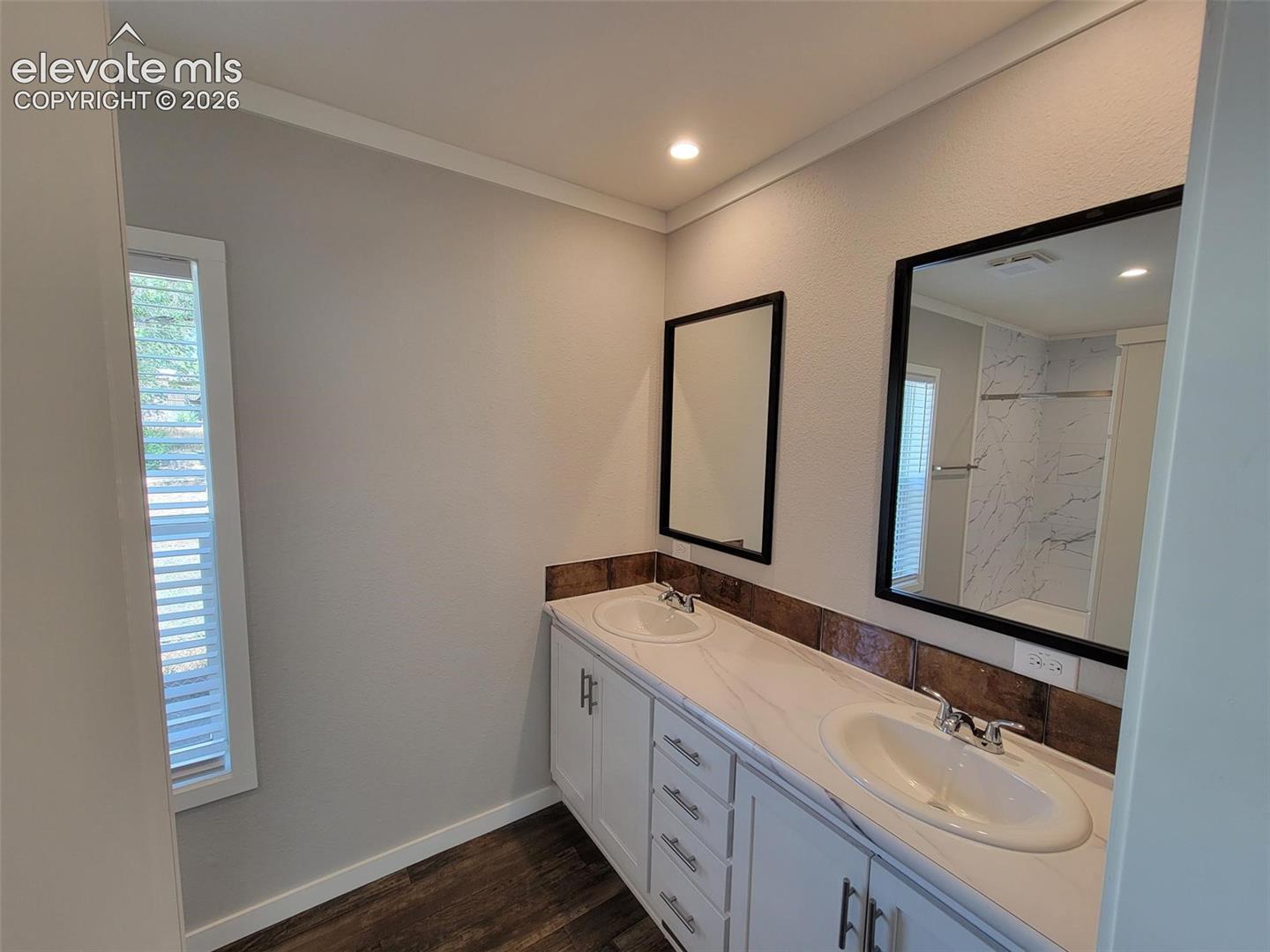 Dual-sink vanity with stone-finish countertop, white cabinetry with metal hardware, tiled backsplash, and dual black-framed wall mirrors