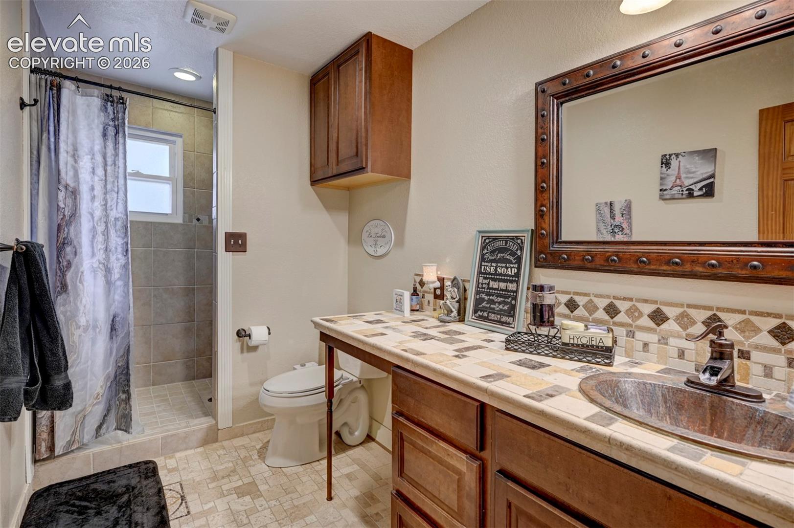 Tile-surround shower with window, integrated recessed lighting, wood-finish vanity with built-in cabinetry, tile countertop, and a decorative framed mirror