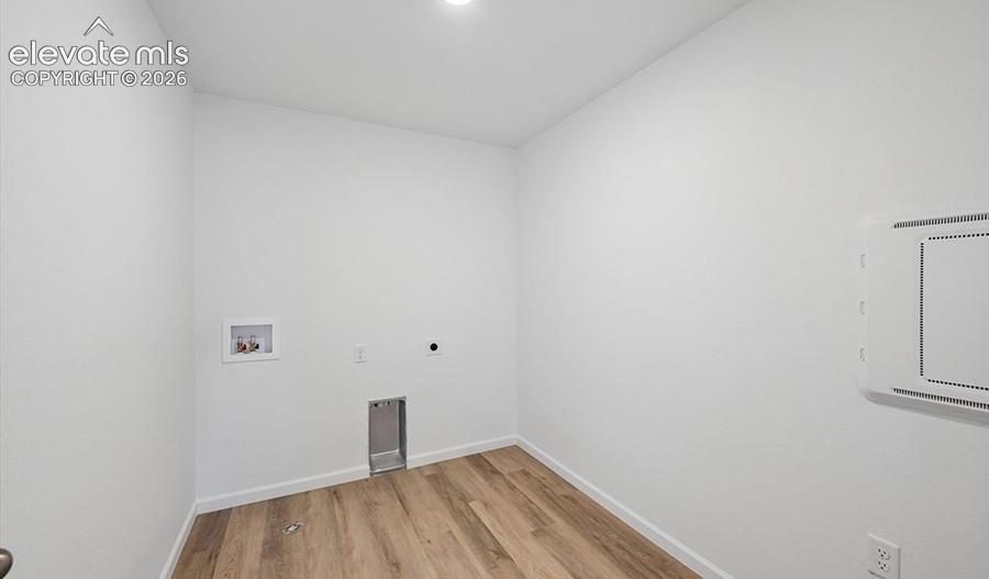 Dedicated utility area featuring wood-finish flooring, recessed water supply valves, drain plumbing, and a wall-mounted electrical panel