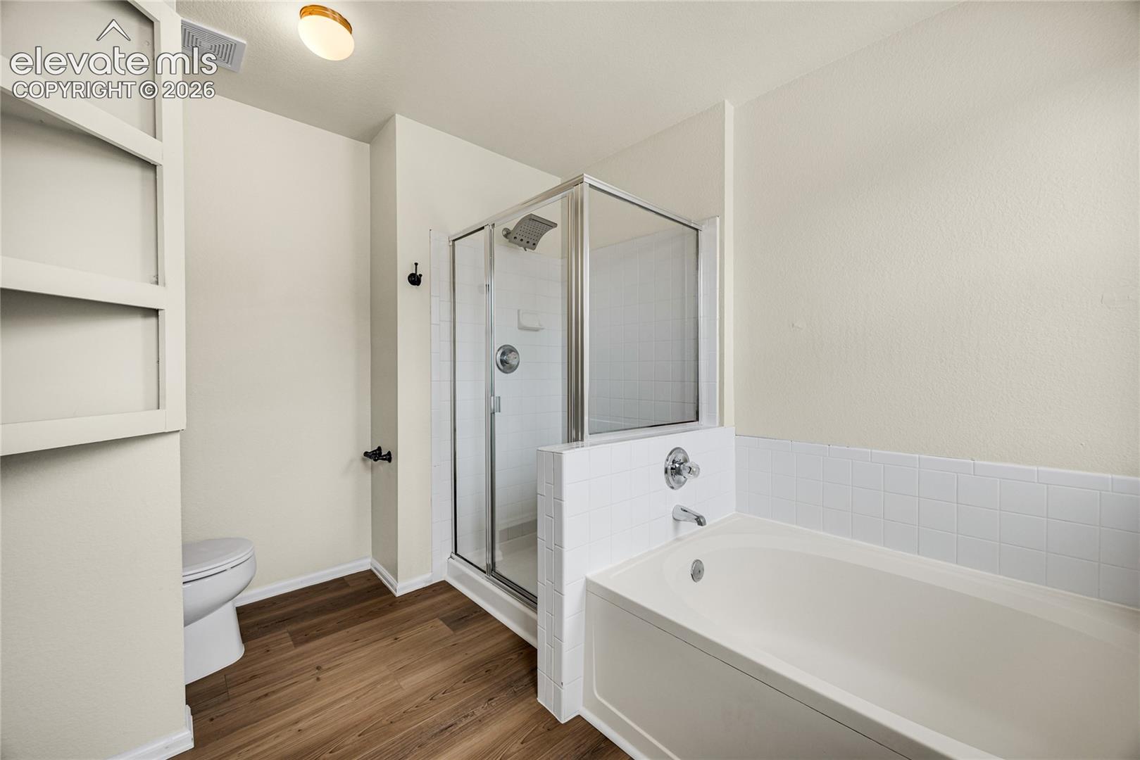 Bathroom featuring a deep soaking tub with white tile surround, glass-enclosed shower, wood-finish flooring, and built-in shelving