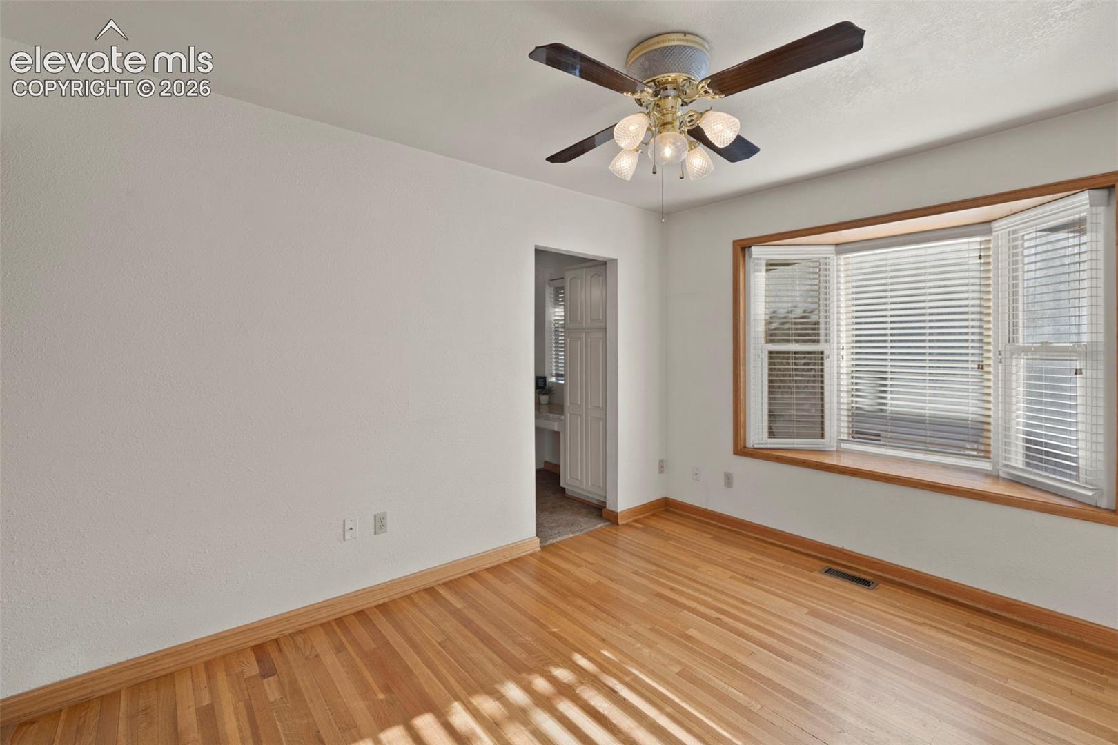 Hardwood flooring, bay window with plantation shutters, ceiling fan with integrated lighting, and white wall finishes