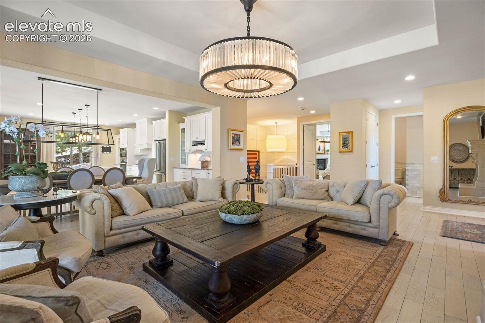 Spacious living area featuring a large central chandelier, wood-finish flooring, and a broad entry point into an adjacent dining and kitchen area