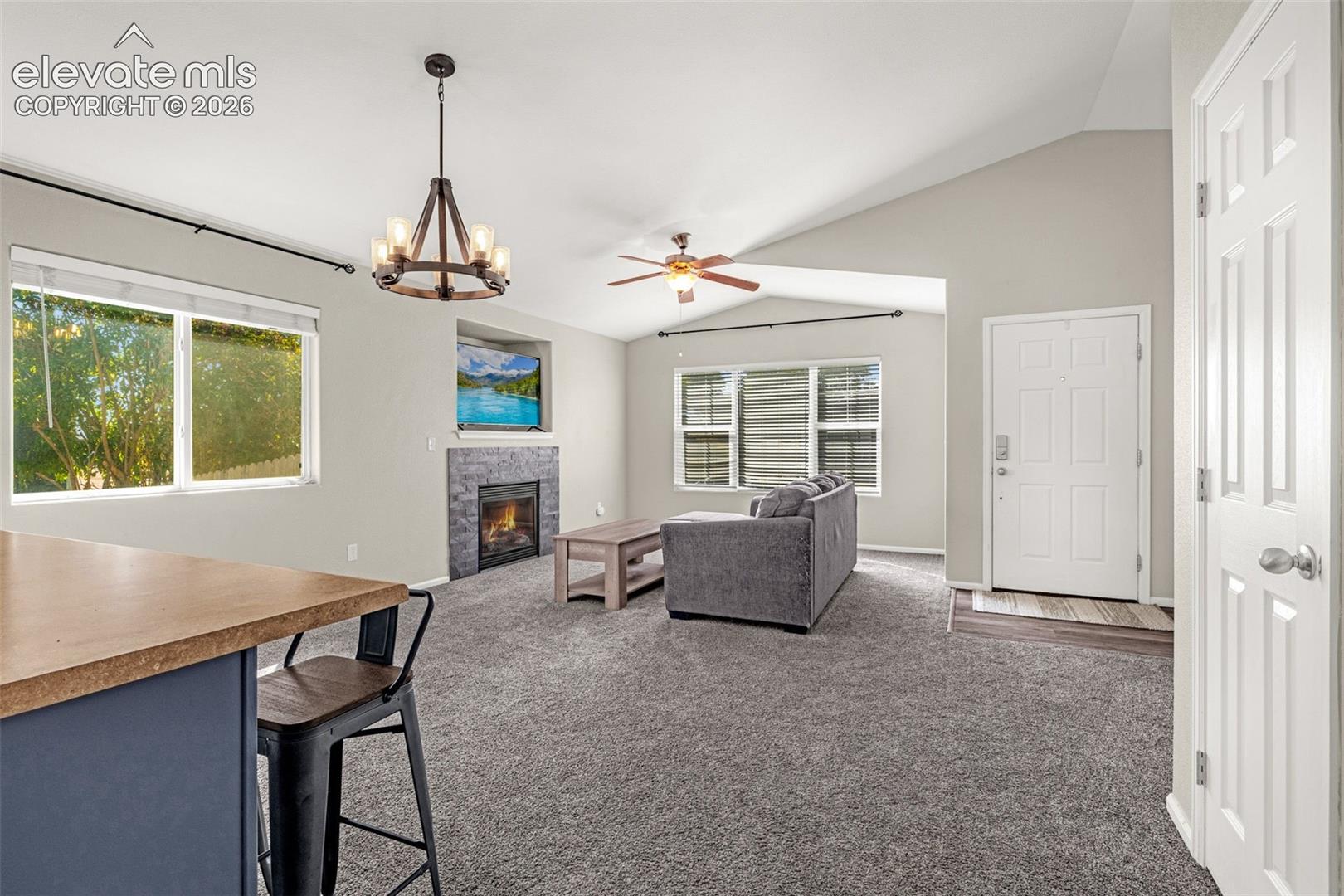 Vaulted ceiling with an overhead fan, stone-clad fireplace, built-in display nook, and wood-finish breakfast bar