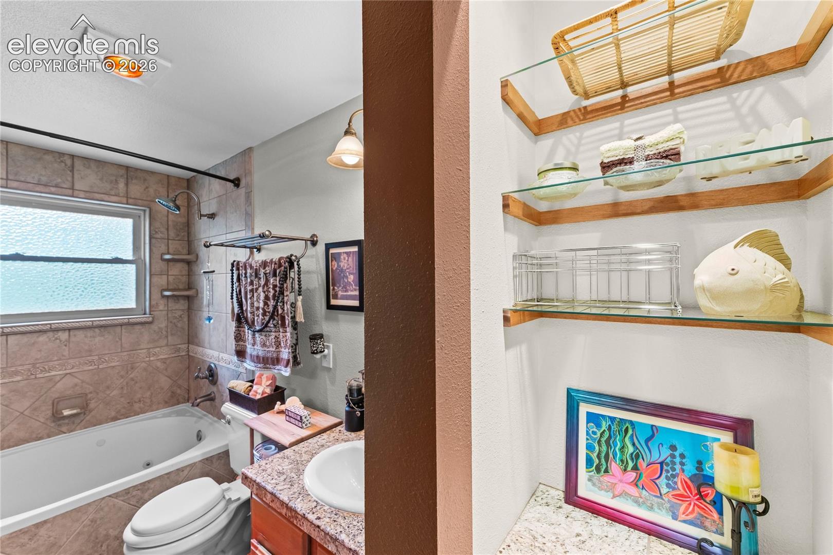 Main bathroom featuring a soaking tub with tiled surround, a window with privacy glass, a vanity with a granite-look countertop and integrated sink, a wall-mounted light fixture, and built-in shelving with glass shelves