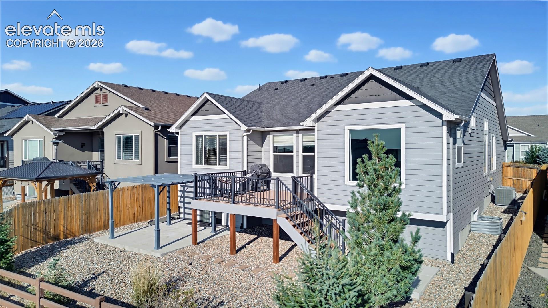 Rear exterior featuring horizontal lap siding, elevated deck with black metal railing, ground-level concrete patio with pergola, and fenced yard with decorative stone ground cover