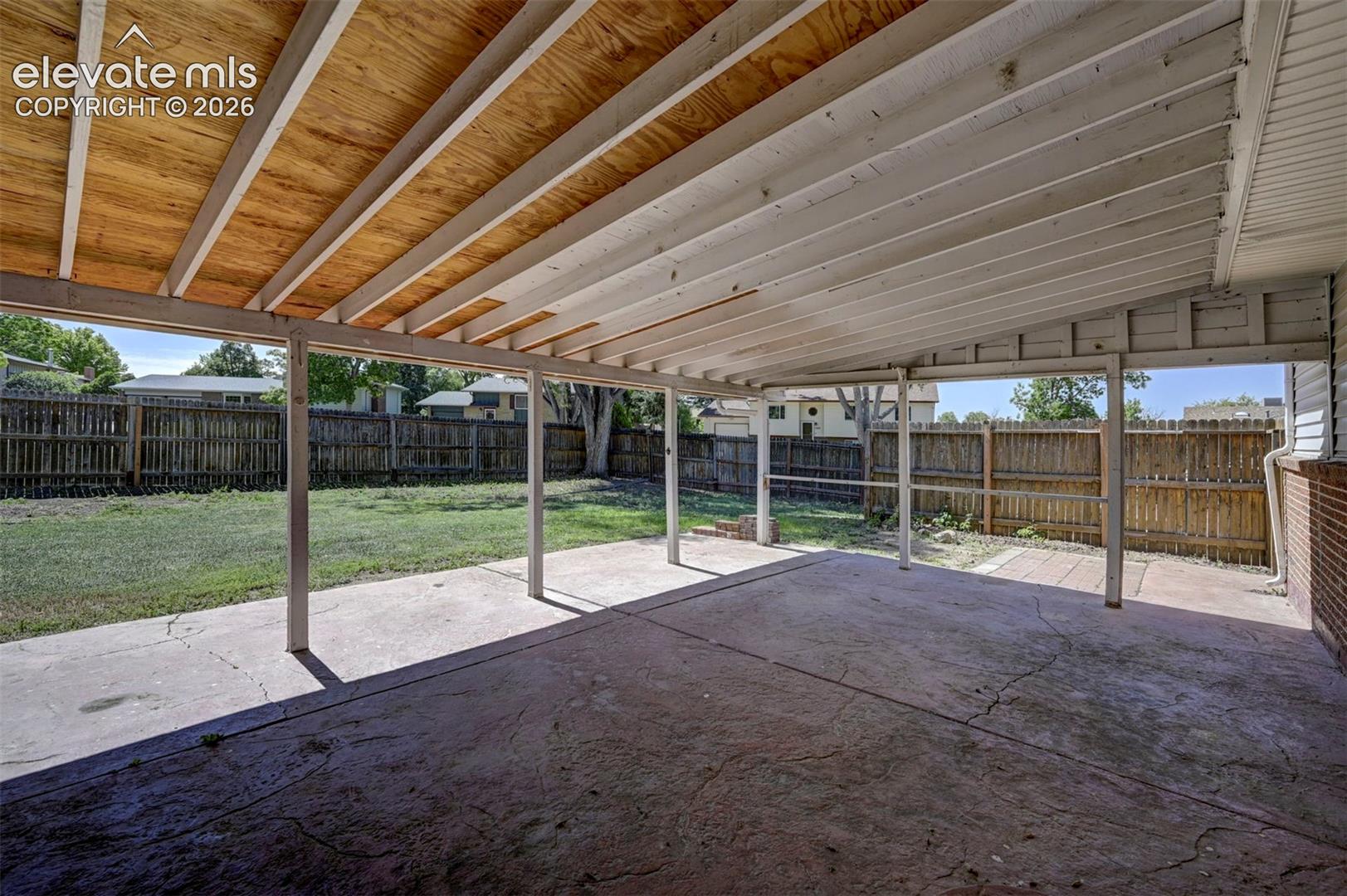 Covered outdoor patio featuring a concrete floor, exposed wooden rafters, and a wood privacy fence enclosure