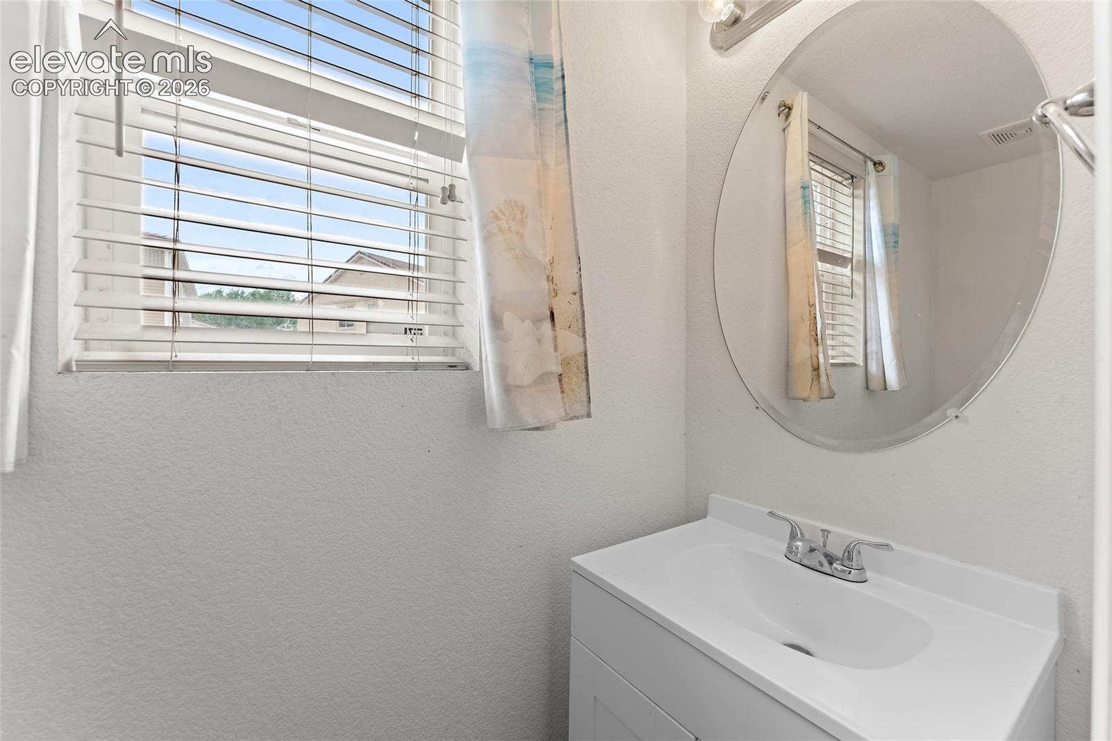 White vanity with integrated sink, chrome double-handle faucet, circular wall-mounted mirror, multi-light vanity fixture, and horizontal window blinds