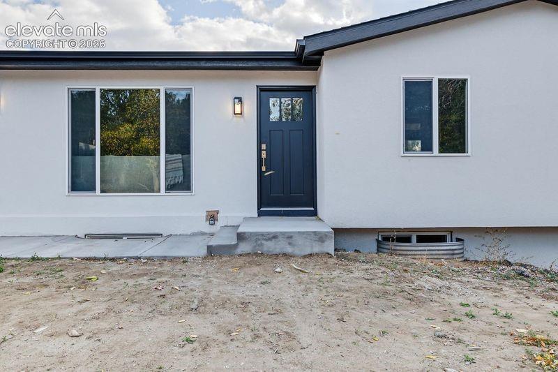 Exterior facade featuring smooth white stucco siding, a dark-toned entry door with inset glass paneling, black roof trim, and large windows