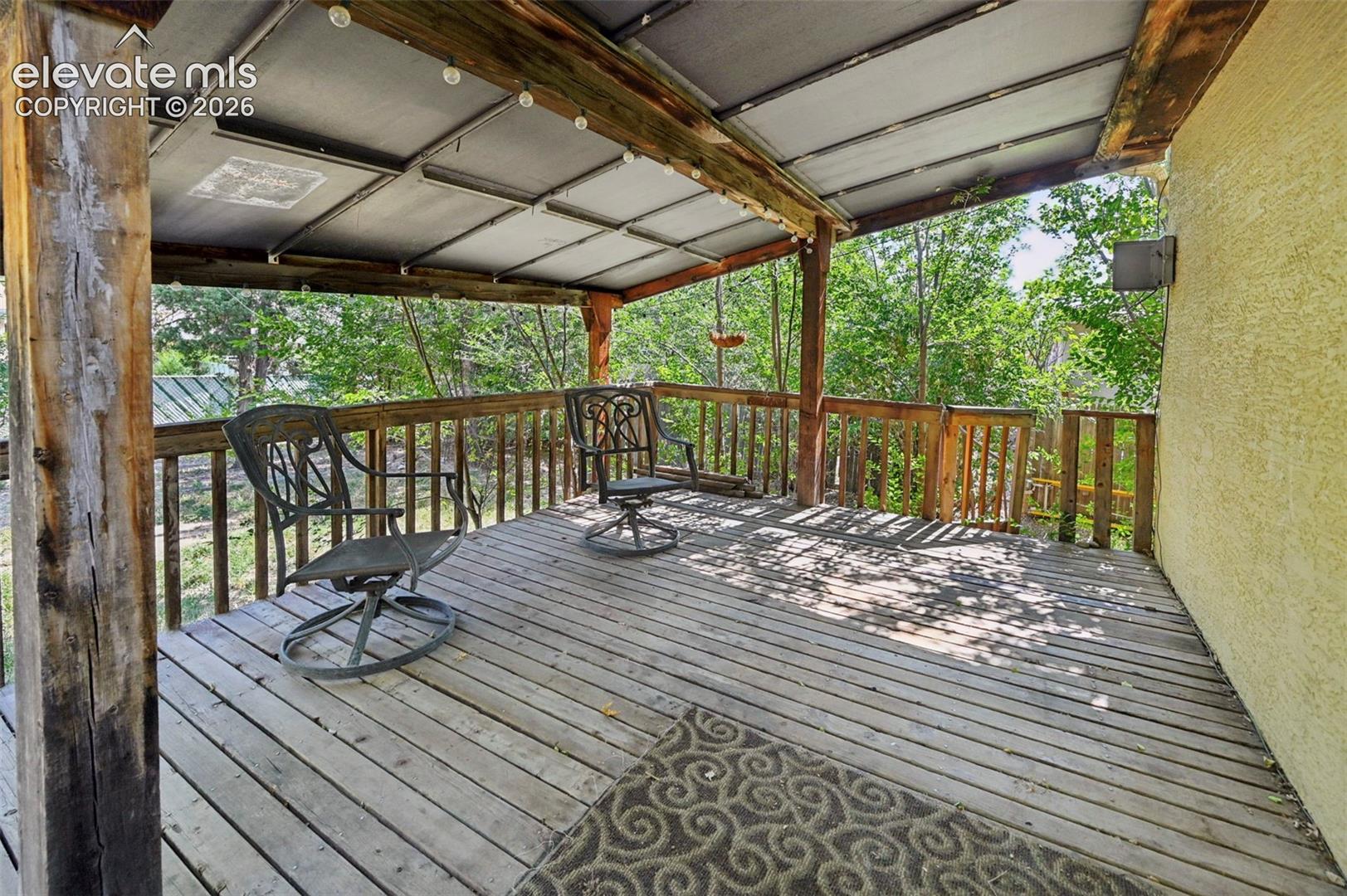 Spacious outdoor deck featuring natural wood flooring and railing, exposed wood beams, a covered patio area, and surrounding mature trees
