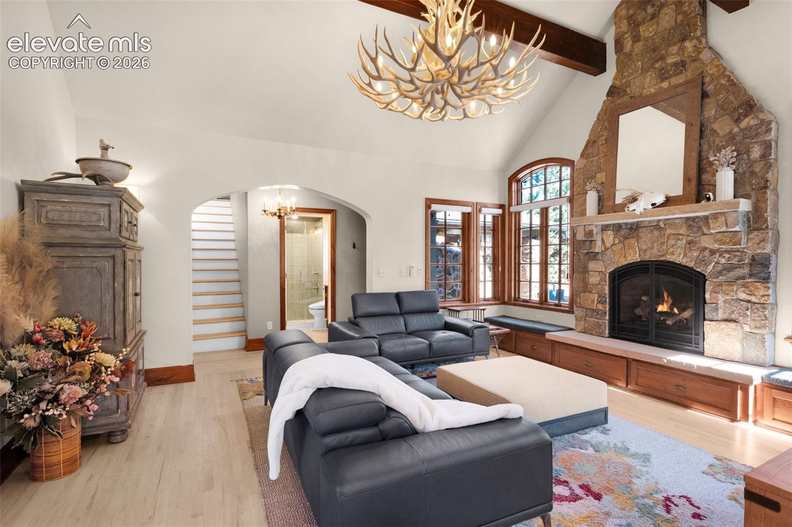 Vaulted ceilings with exposed wood beams, floor-to-ceiling stone fireplace with a wood mantel, wood-finish flooring, and arched entryway