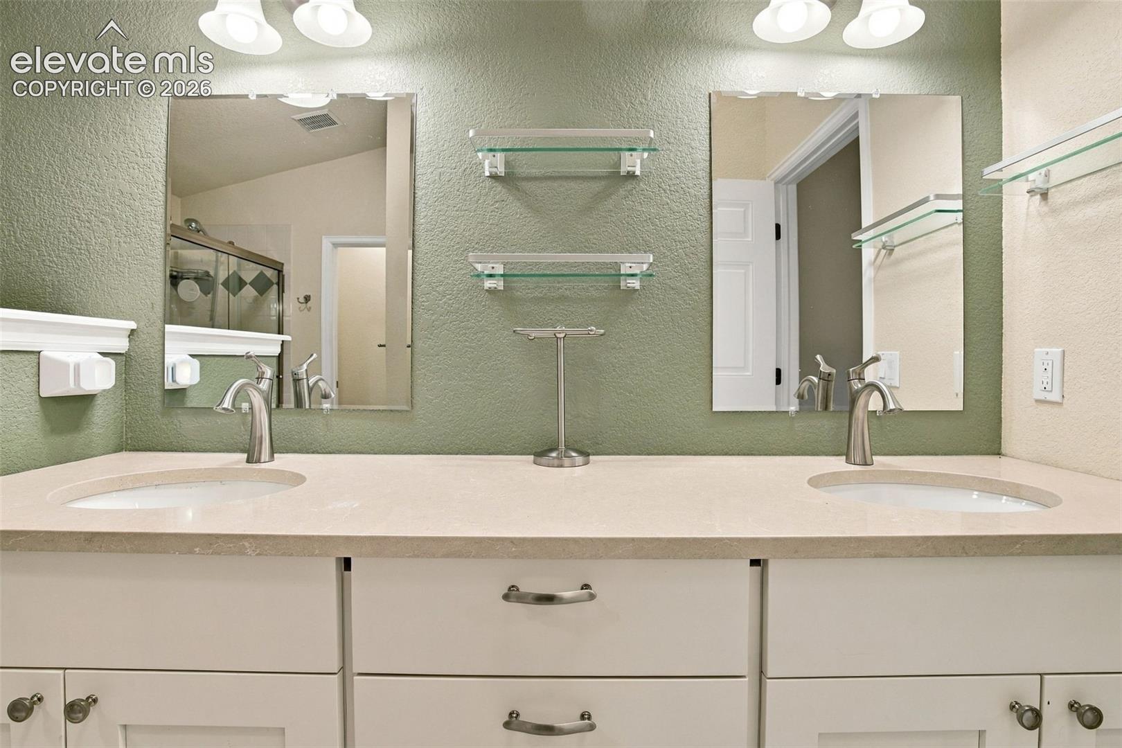 Double-sink vanity featuring a neutral solid-surface countertop, white shaker-style cabinetry with brushed nickel hardware, dual wall-mounted mirrors, and glass shelving