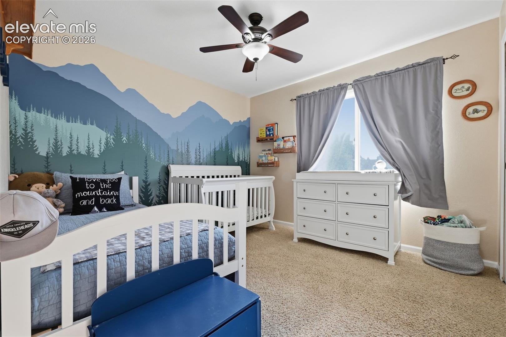 Spacious room featuring neutral carpeting, a dual-pane window with drapery, a ceiling fan with integrated lighting, and a painted mountain mural
