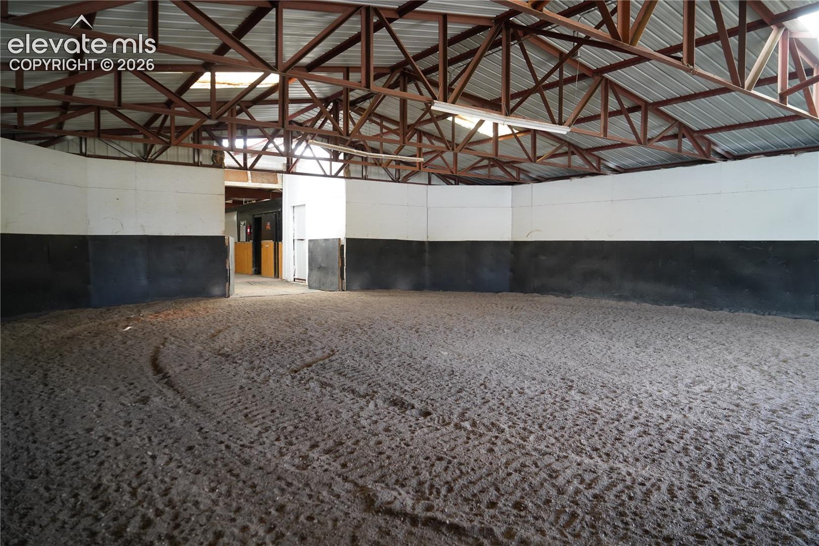 Expansive indoor arena with a sand footing, high ceilings with exposed steel beams, and multiple overhead light fixtures