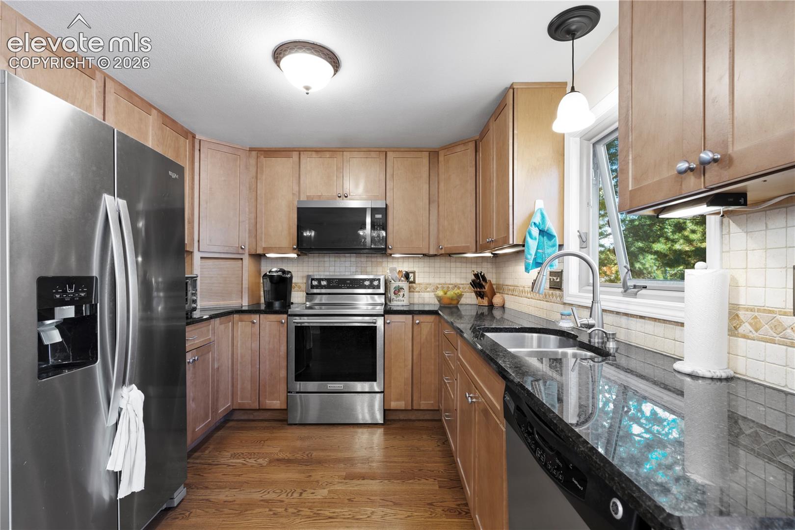 Kitchen featuring wood-finish flooring, stainless steel appliances, dark countertops, wood cabinetry, and a tile backsplash