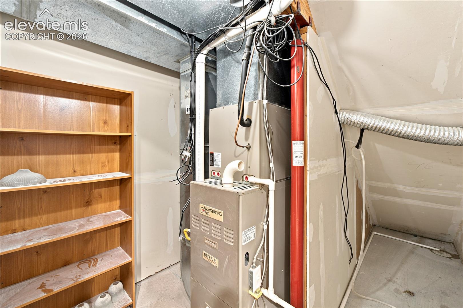 Utility room featuring a furnace unit, exposed ductwork, flexible ventilation tubing, and a built-in wood-finish shelving unit