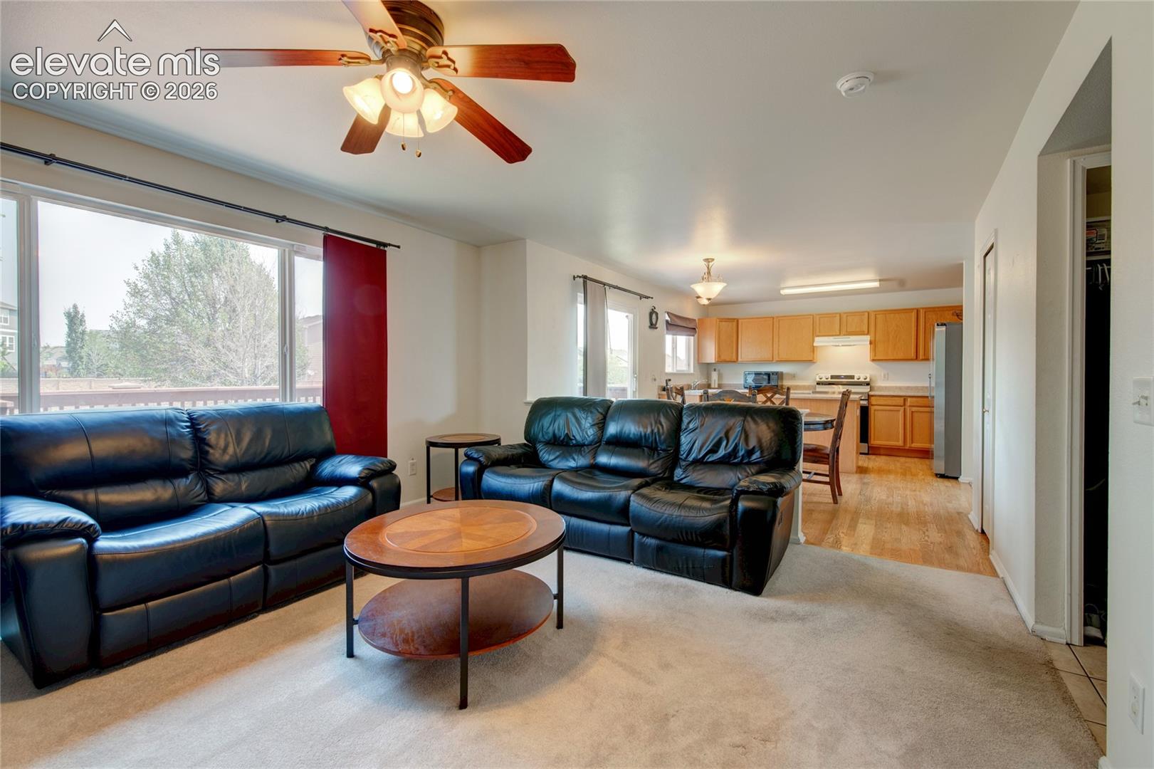 Open-concept living area featuring a large window, a ceiling fan with integrated lighting, and a kitchen visible in the background
