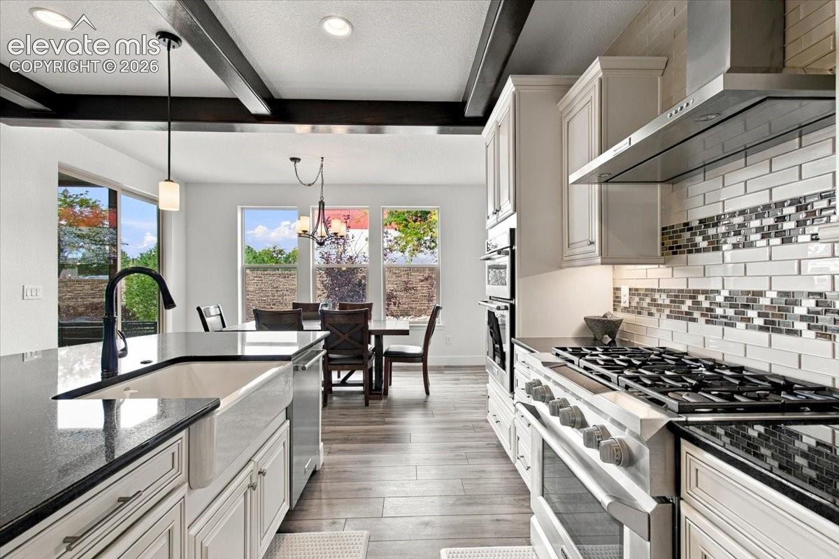 Kitchen featuring wood-finish flooring, exposed dark ceiling beams, shaker-style cabinetry, farmhouse sink with matte black faucet, and stone-tiled backsplash