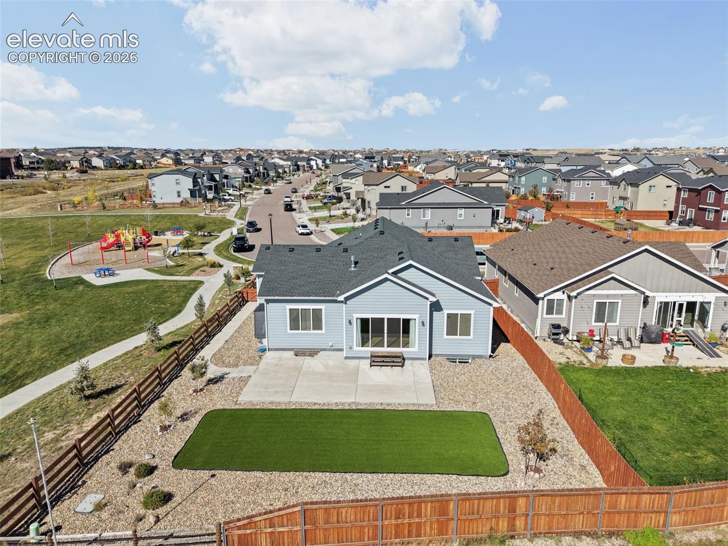 Single-story residence featuring light-toned horizontal siding, dark-toned roof shingles, a concrete patio, artificial turf lawn, and perimeter wood fencing