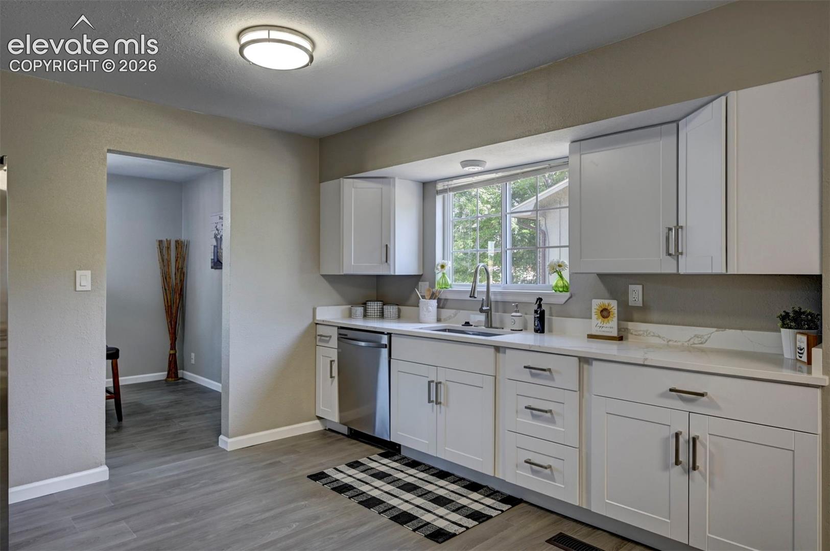 Kitchen featuring white Shaker-style cabinetry, light-toned stone-finish countertops, stainless steel dishwasher, undermount sink with a gooseneck faucet, and wood-finish flooring