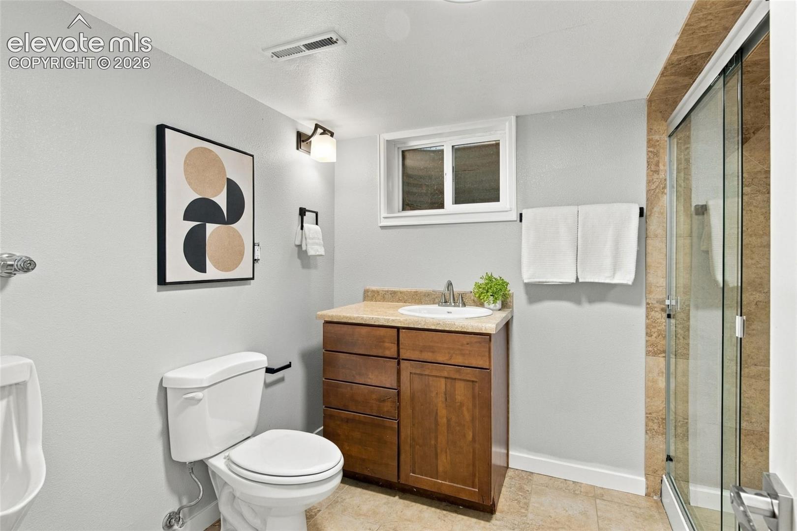 Bathroom featuring a wood-tone vanity with a solid-surface countertop, an integrated sink, a glass-enclosed shower with stone-tile surround, and a wall-mounted light fixture