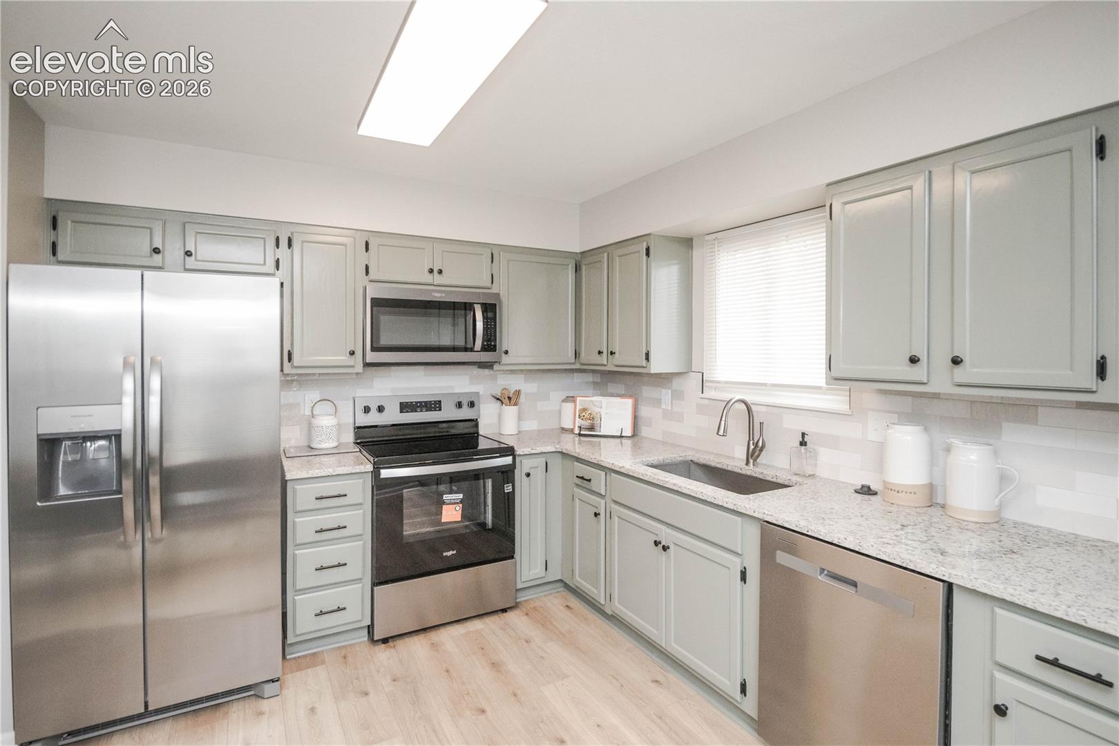Kitchen featuring sage green cabinetry, light-toned stone-finish countertops, stainless steel appliances, a tiled backsplash, and wood-finish flooring
