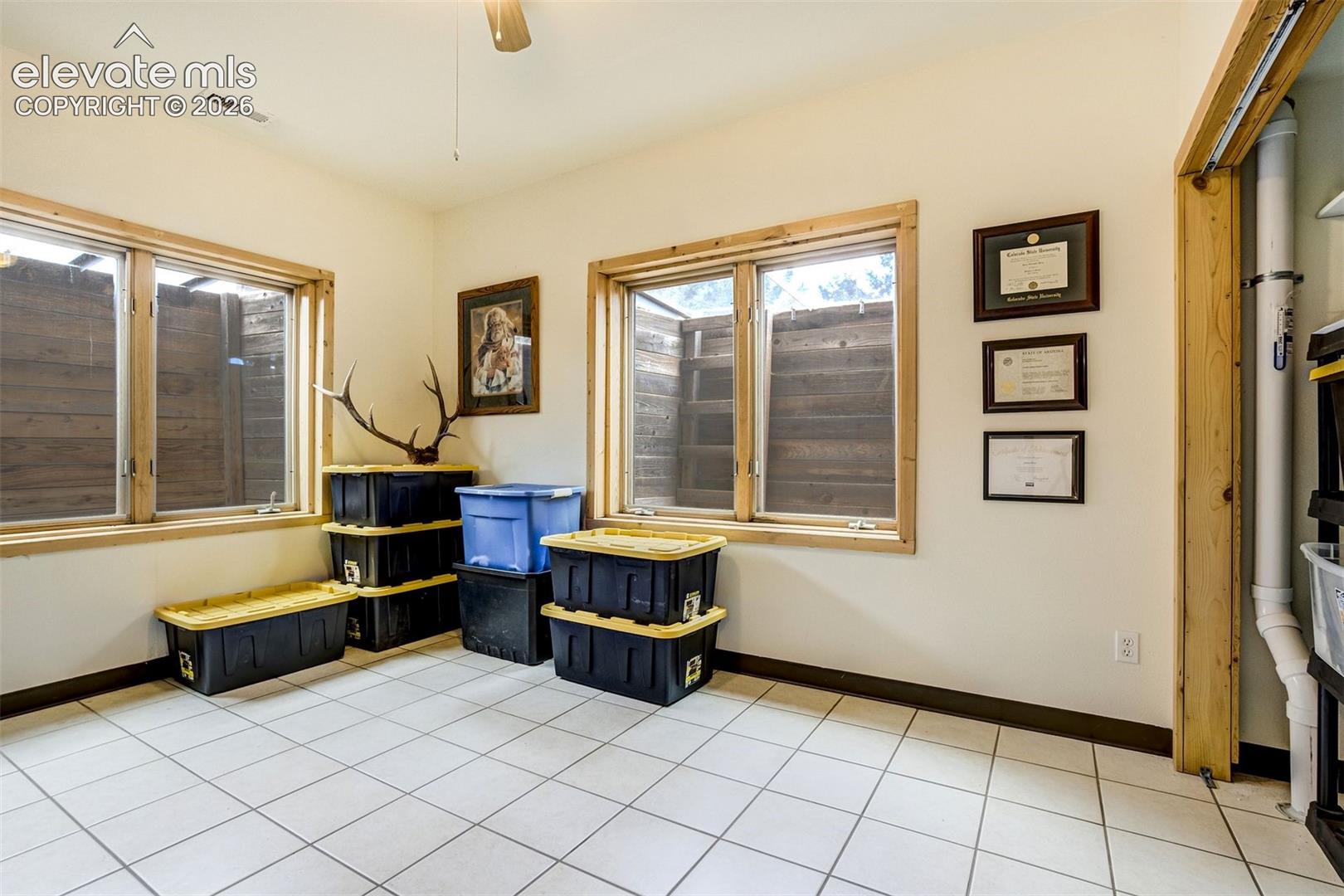 Room featuring white tile flooring, two windows with natural wood trim, white painted walls, and a ceiling fan