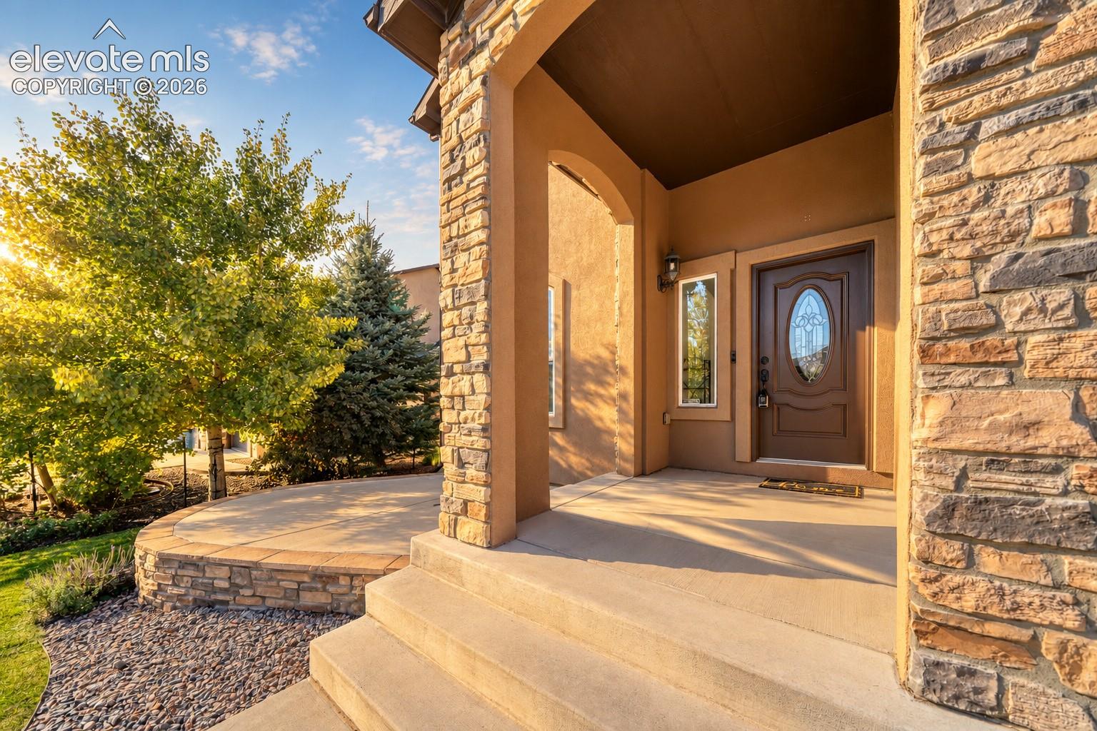 Stone and stucco exterior detailing, covered entry, solid wood-finish entry door with oval glass insert, concrete front steps, and a curved concrete patio with stone retaining wall
