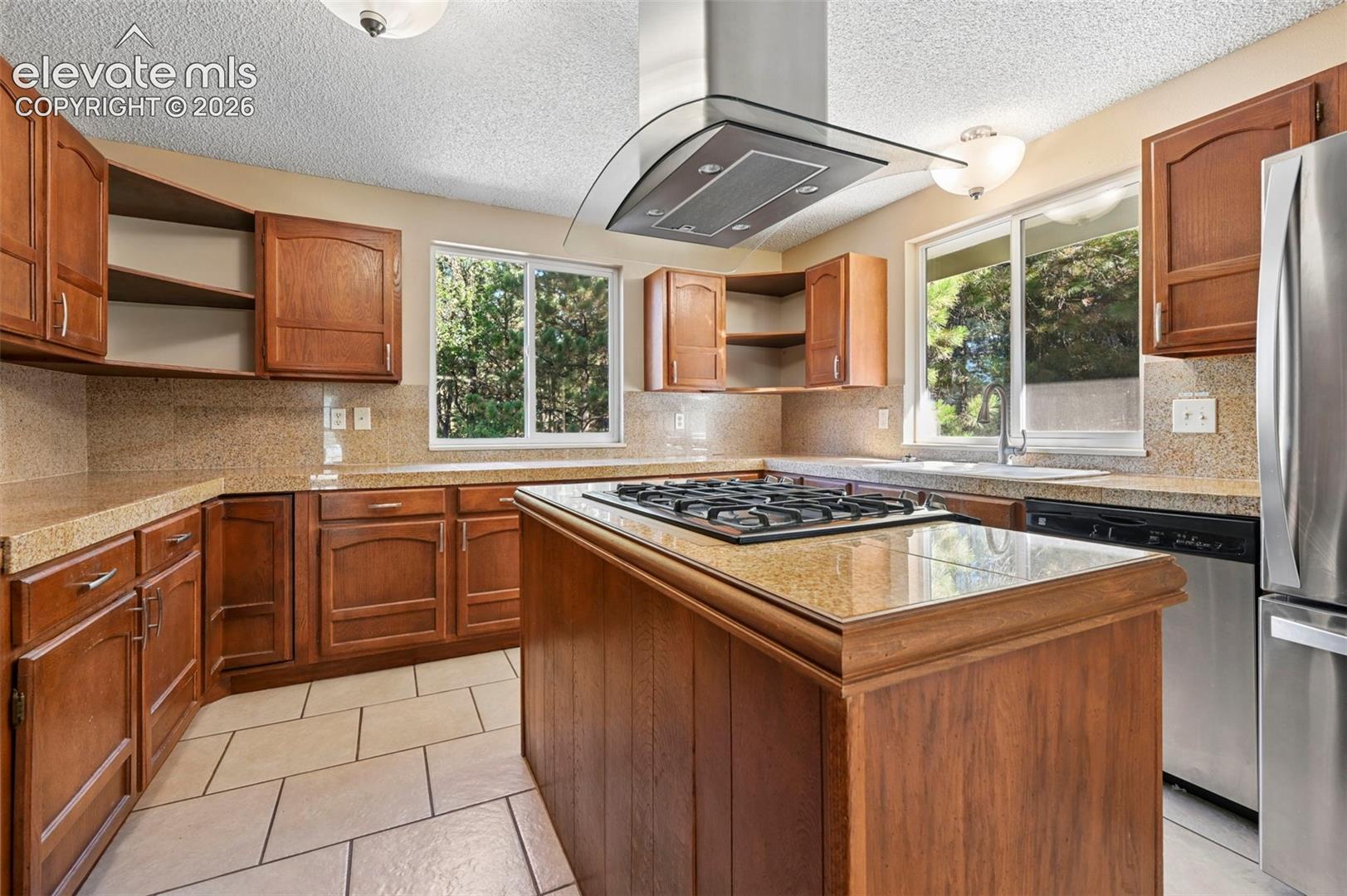 Kitchen featuring wood cabinetry, solid-surface countertops, center island with an integrated gas cooktop, stainless steel ventilation hood, and tile flooring
