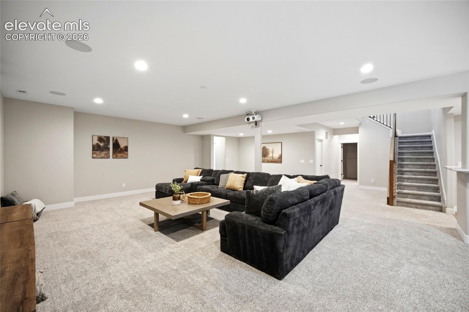 Open-concept room featuring neutral wall-to-wall carpeting, recessed ceiling lighting, integrated ceiling speakers, and a carpeted staircase with a wood handrail