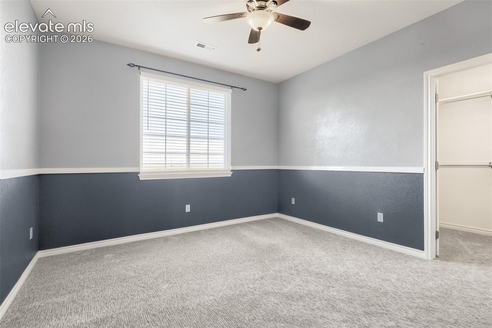 Inviting second-floor bedroom showcasing tasteful two-tone accent paint accented by decorative chair rail trim. Includes plush wall-to-wall carpeting, a ceiling fan, a bright window with blinds, and a private closet with built-in storage.