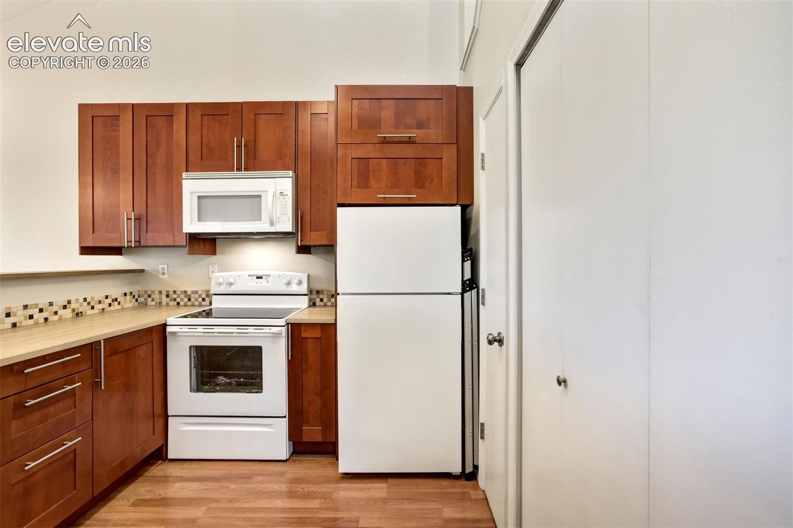 Kitchen featuring wood-finish cabinetry, neutral-toned countertops, a multi-colored tile backsplash, white appliances, and wood-finish flooring
