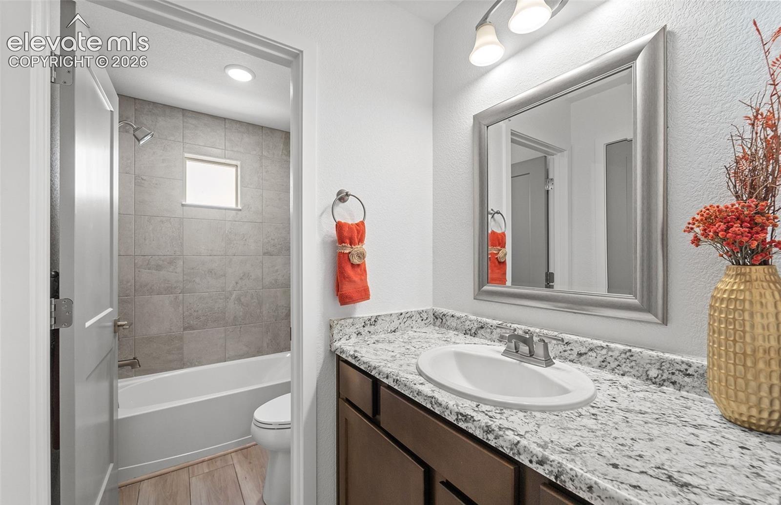 Bathroom featuring a stone-patterned vanity countertop, integrated sink, framed wall mirror, and a tub with large-format tile surround